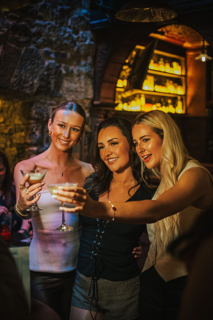 Three women smiling and raising cocktails for a photo in a warmly lit pub setting, with shelves of spirits glowing behind them and stone walls adding to the cosy atmosphere.