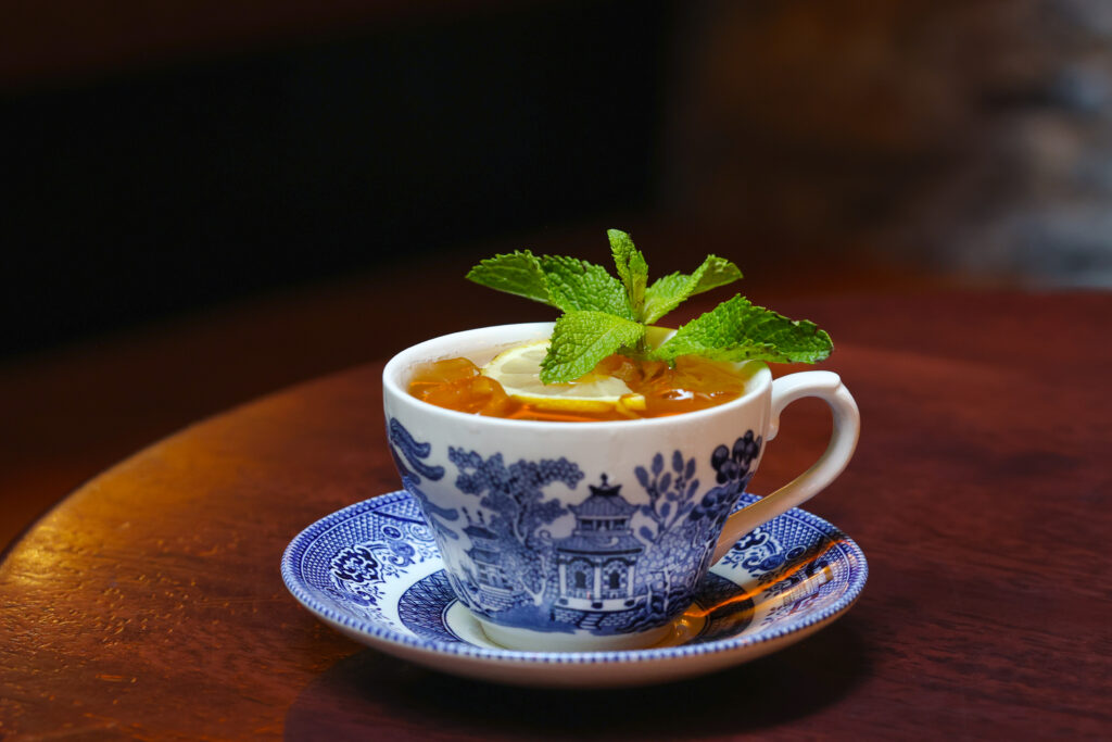 The Poet’s Measure cocktail served in a vintage blue and white teacup on a matching saucer, garnished with lemon slices and fresh mint.
