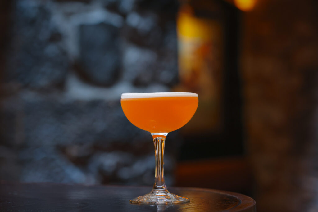High Kings and Queens cocktail served in a coupe glass, featuring a rich amber-orange hue with a light frothy top, placed on a dark wooden surface with a blurred background