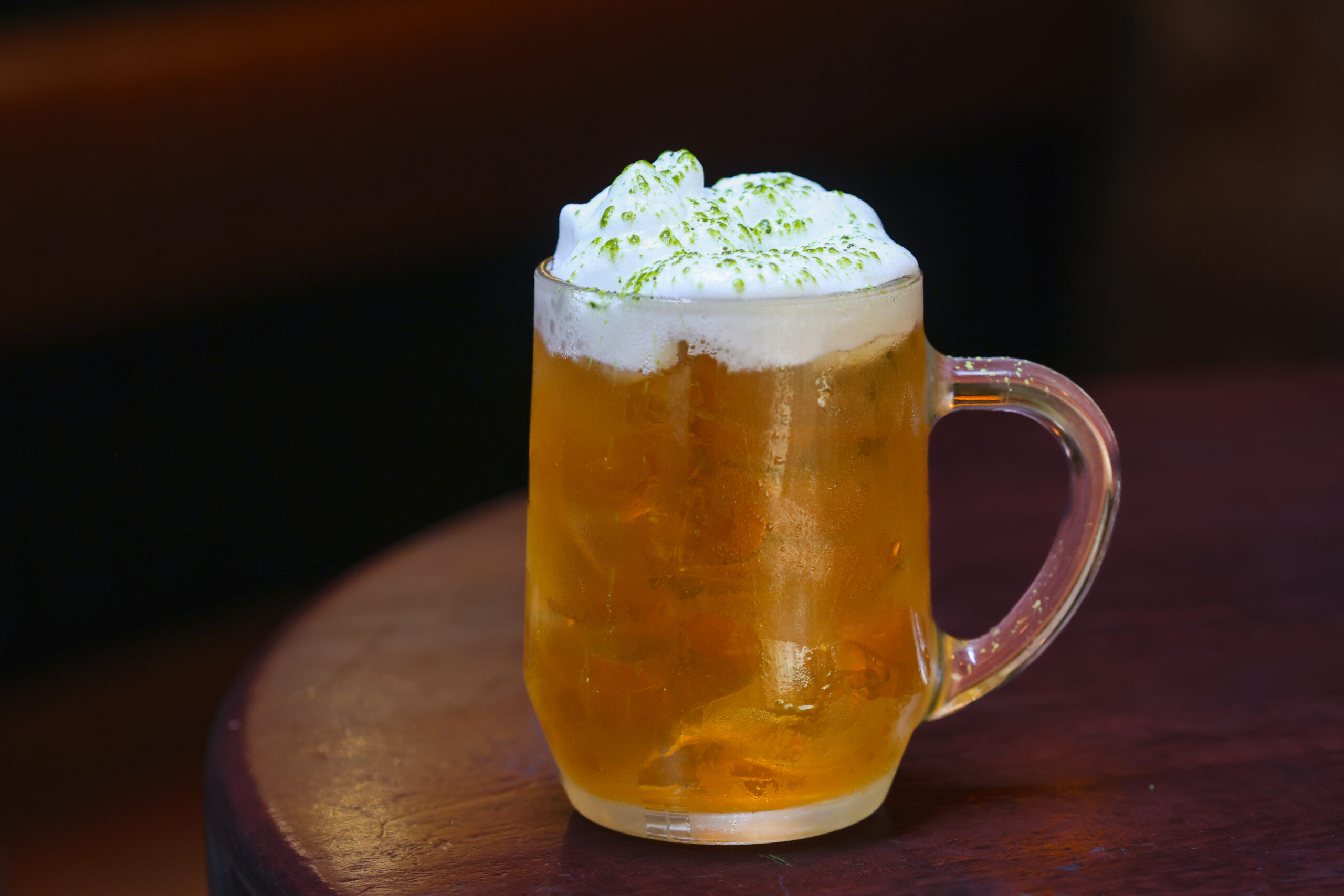 The Hall of Fame cocktail served in a clear glass tankard over ice, topped with a generous layer of tropical foam and a sprinkle of green powder, set on a dark wooden surface.