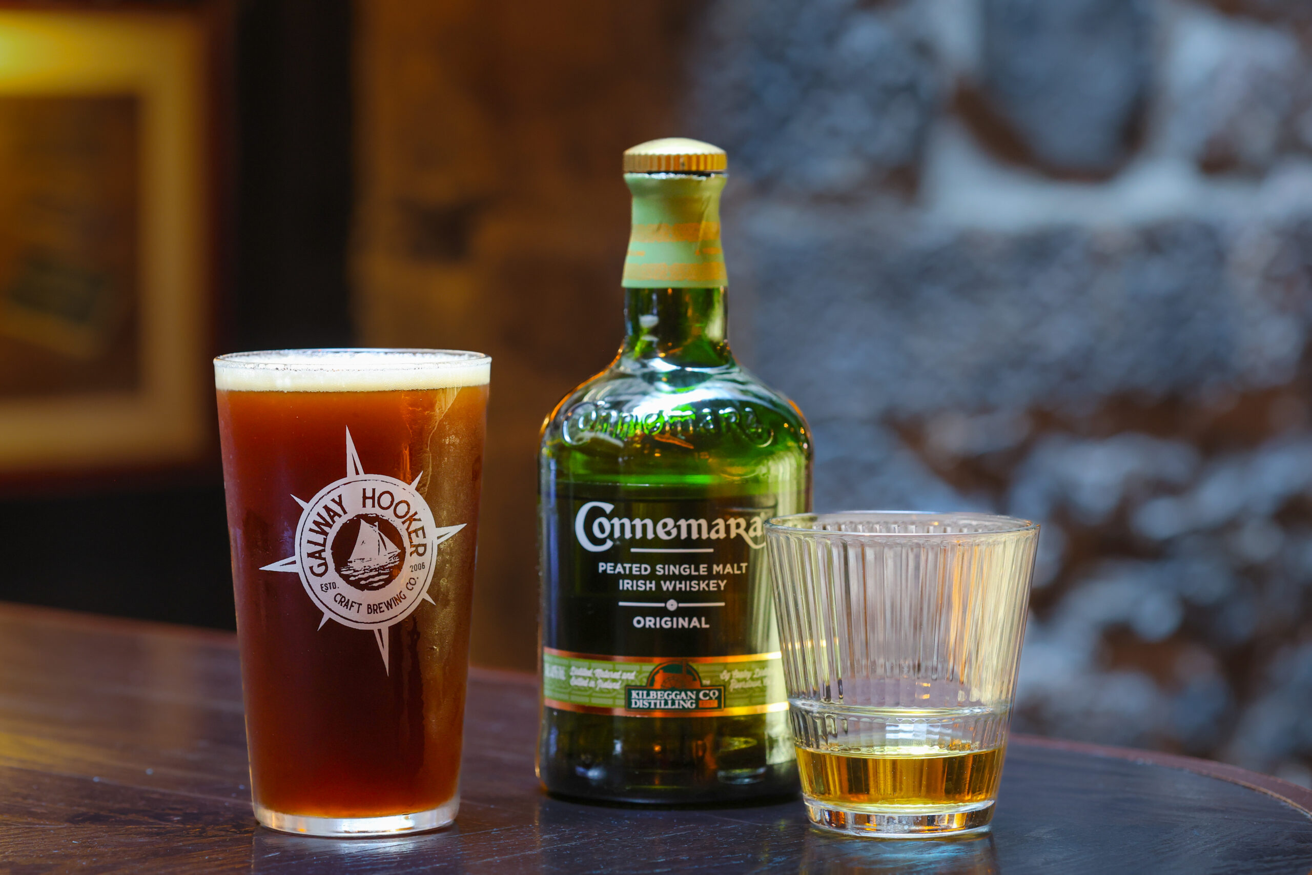 Boilermaker set featuring a pint of Galway Hooker beer, a bottle of Connemara Peated Single Malt Irish Whiskey, and a glass of whiskey, all arranged on a dark wooden bar.